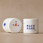 Two white opaque jars of Blue Sage THC product on a neutral background, with one upright showing the blue "BLUE SAGE THC" text logo and the other resting on its side showing an orange fruit and star graphic on its cap.