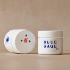 Two white opaque jars of Blue Sage THC product on a neutral background, with one upright showing the blue "BLUE SAGE THC" text logo and the other resting on its side showing an orange fruit and star graphic on its cap.